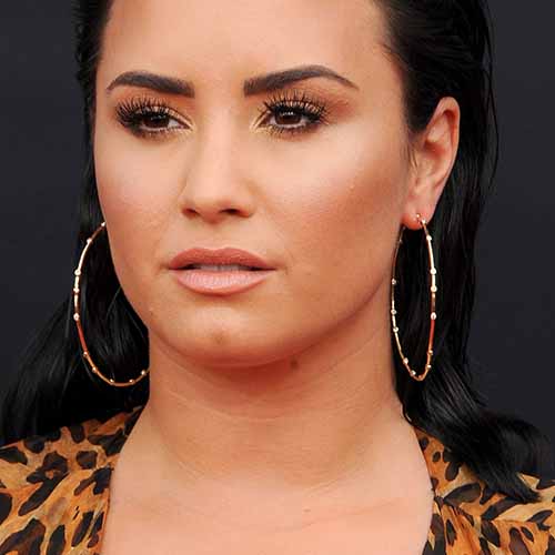 Singer Demi Lovato in 2018, with leopard-print dress, shoulder-length black hair, and gold hoop earrings. 